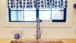 A kitchen with a stainless steel sink, a window with a checkered valance, and a sign above the window.