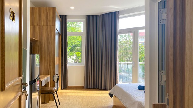 A modern hotel room with a wooden wardrobe, a bed with a white duvet, a desk with a chair, and a large window with curtains.