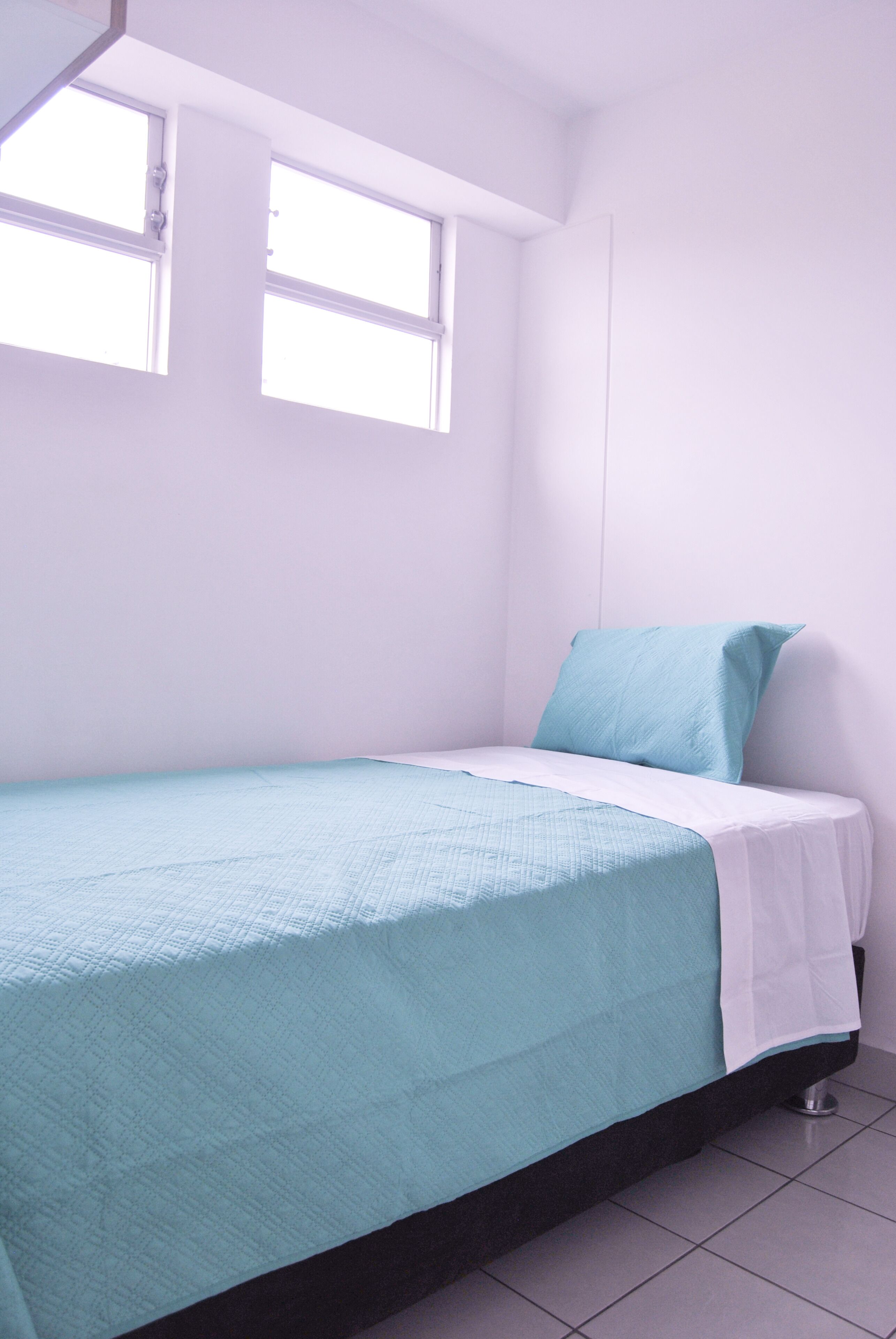 Una habitación pequeña con cama individual, ventana, paredes blancas y colcha color turquesa.