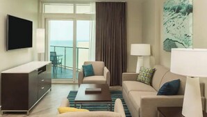 Une chambre d’hôtel équipée d’un canapé, d’un fauteuil, d’un téléviseur à écran plat, d’un bureau et offrant une vue sur la mer grâce à une porte coulissante.