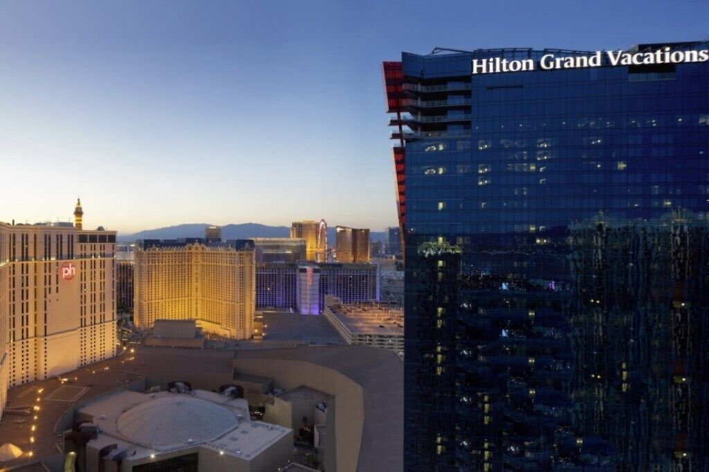 A modern hotel with a prominent name sign, a rooftop area, and surrounding high-rise buildings.