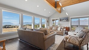 A spacious living room with large windows offering mountain views, a high ceiling with exposed beams, and a mix of leather and patterned furniture. - Stunning views, sleeps 10, firepit, decks, hot tub (Heber City)