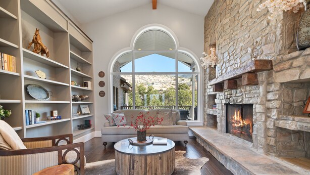 A spacious living room with a stone fireplace, a large window overlooking a scenic landscape, and a cozy seating area with a round coffee table and bookshelves. - Stunning views, sleeps 10, firepit, decks, hot tub (Heber City)