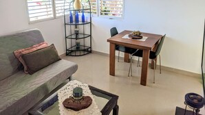A living room with a sofa, a dining table with chairs, a coffee table, and a window with blue curtains. - Nice & Cosy Apartm. for max. 4 Guests next to Pool (Sosua)