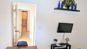 A room with a wooden dining table, a television mounted on the wall, a small side table with a lamp, and a potted plant on a shelf. - Nice & Cosy Apartm. for max. 4 Guests next to Pool (Sosua)