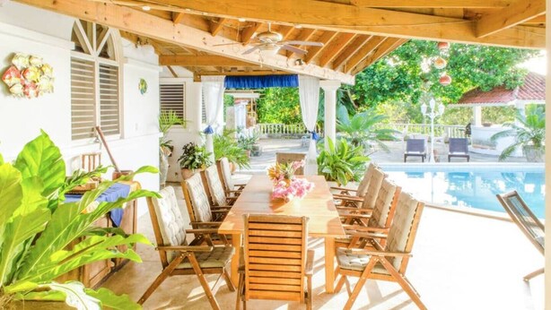 An outdoor patio with a wooden table and chairs, surrounded by potted plants and a view of a swimming pool. - Nice & Cosy Apartm. for max. 4 Guests next to Pool (Sosua)