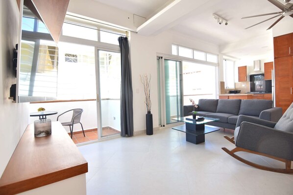 A modern living room with a sofa, a coffee table, and a rocking chair. There is a glass door leading to a balcony. - Bright & Clean 2BR in BEST Colonial Zone Location (Santo Domingo)