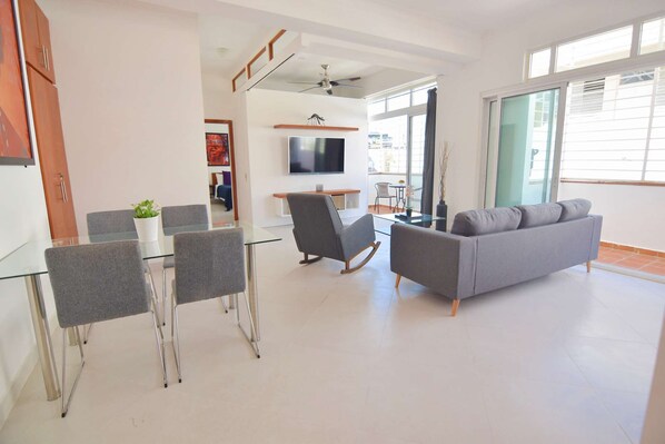 Una sala de estar moderna con un sofá gris, una mesa de comedor de cristal con sillas, un televisor montado en la pared y un balcón con vistas.