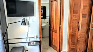 Una habitación con escritorio, televisor montado en la pared, armario de madera y baño visible a través de una puerta abierta.
