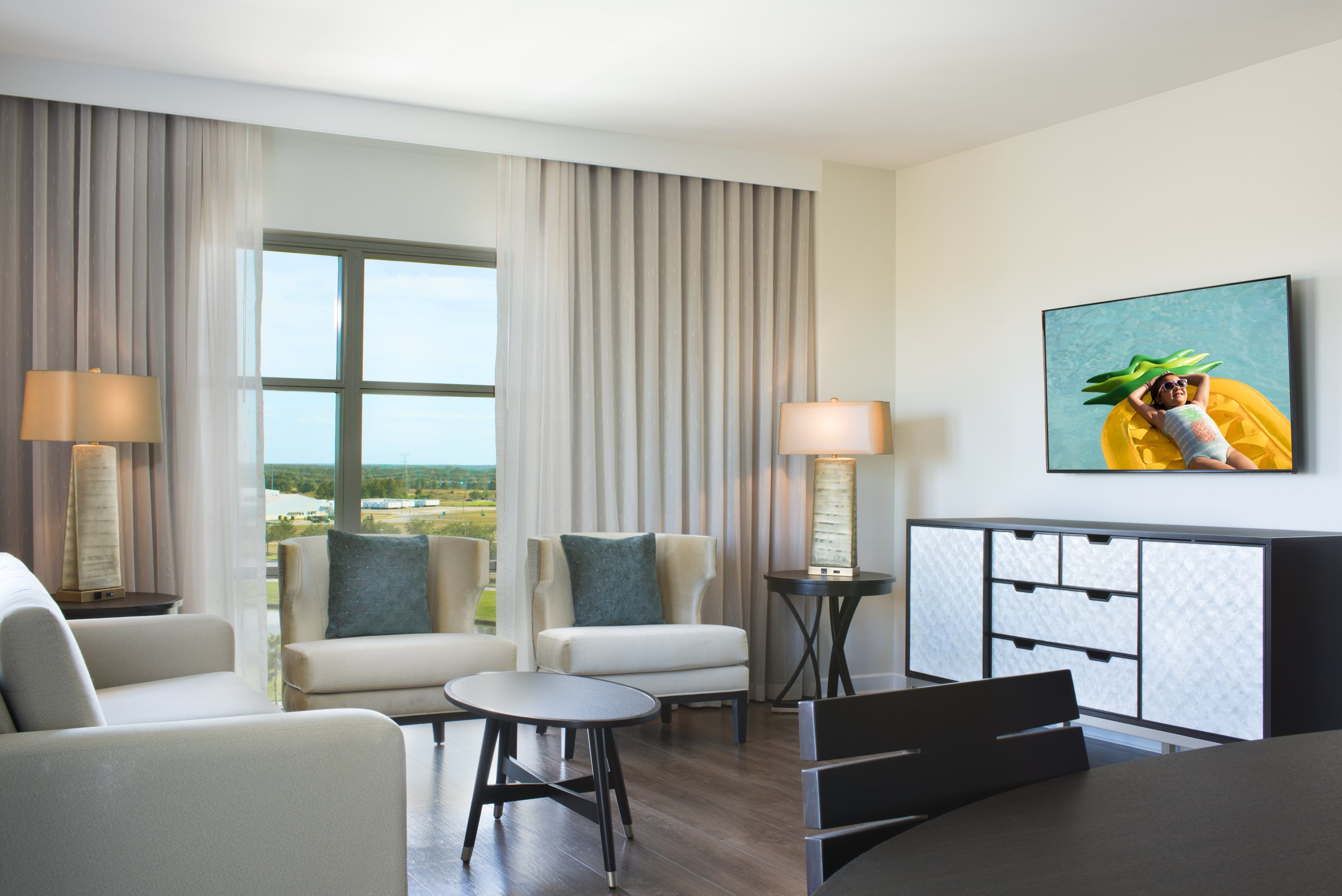 A modern hotel room with a large window, two sofas, a small round table, a cabinet, and two lamps.