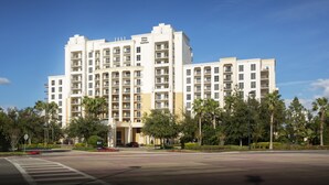 A multi-story hotel with a central entrance, surrounded by palm trees and a clear blue sky.