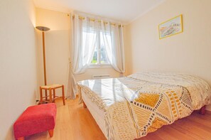 A bedroom with a bed, a chair, a red bench, a floor lamp, and a window with curtains.