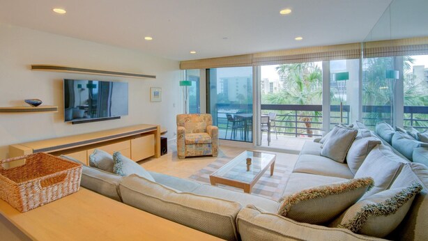 A modern living room with a sofa, a coffee table, a TV, and a balcony with outdoor furniture. - Beachplace 7 - 202 by FGC (Longboat Key)