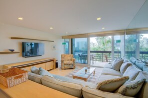 A modern living room with a sofa, a coffee table, a TV, and a balcony with outdoor furniture. - Beachplace 7-202 by FGC (Longboat Key)