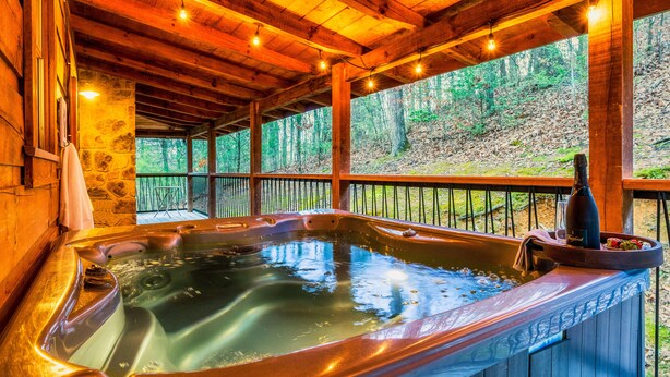 A hot tub with a bottle of wine and a tray of fruit, surrounded by wooden beams and stone walls.