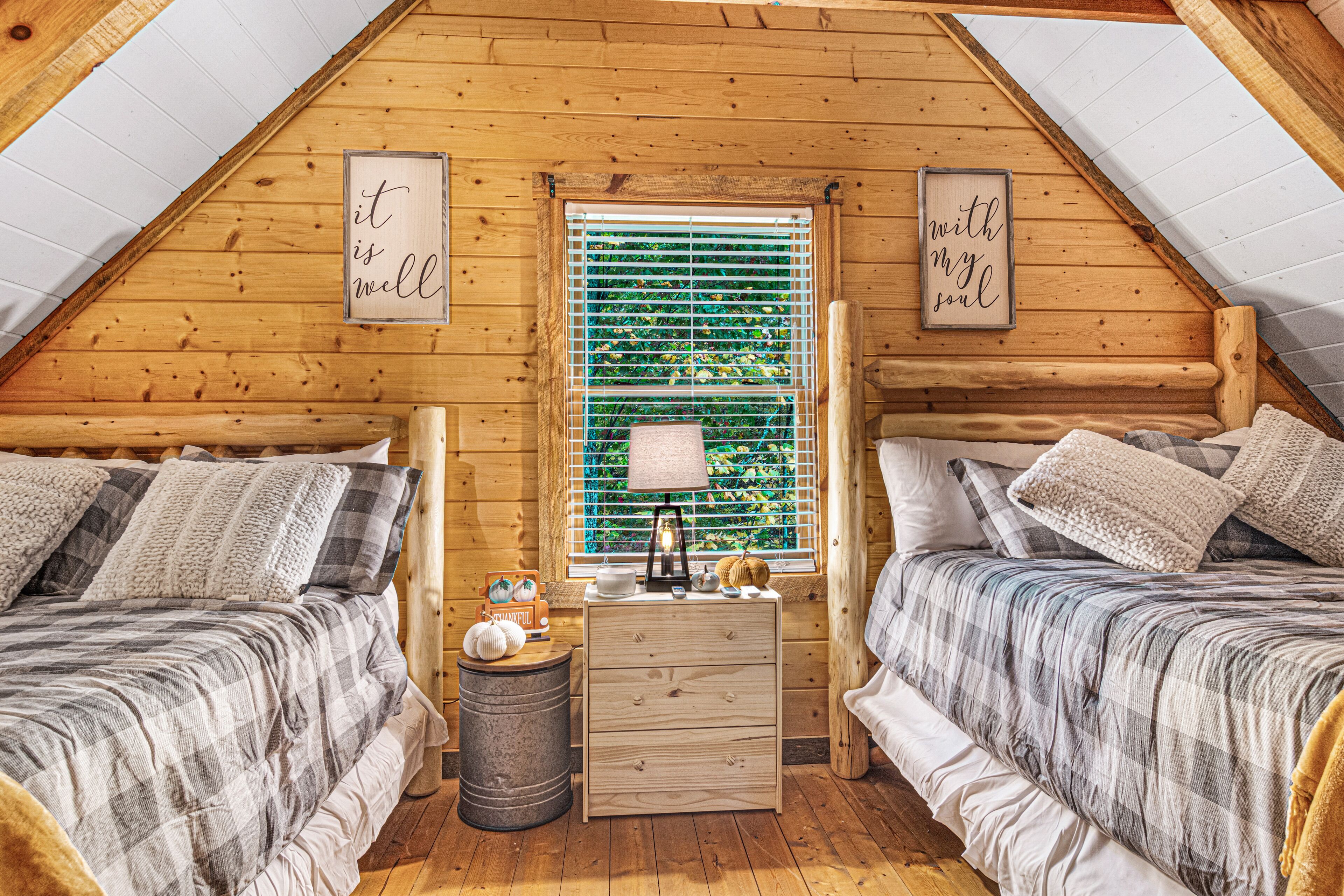 Un acogedor dormitorio en áticos con dos camas, paredes de madera, un ventanal con persianas y una pequeña mesita de noche con lámpara.