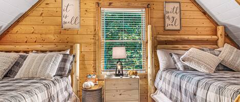 Un acogedor dormitorio en áticos con dos camas, paredes de madera, un ventanal con persianas y una pequeña mesita de noche con lámpara.
