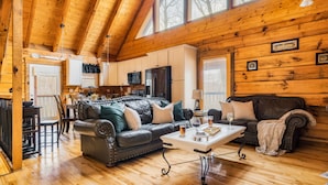 A cozy cabin interior with a wooden ceiling, a dining area, a living room with a sofa and a coffee table, and a kitchen with appliances.