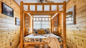 A wooden cabin bedroom with a bed, two stools, a ceiling fan, and framed pictures on the walls.