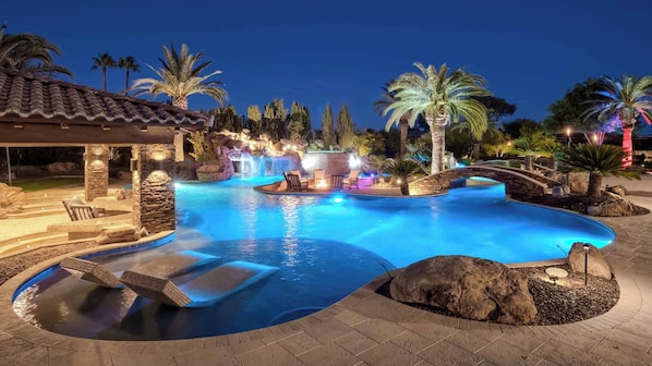 A swimming pool with a waterfall feature, surrounded by palm trees and a stone patio.
