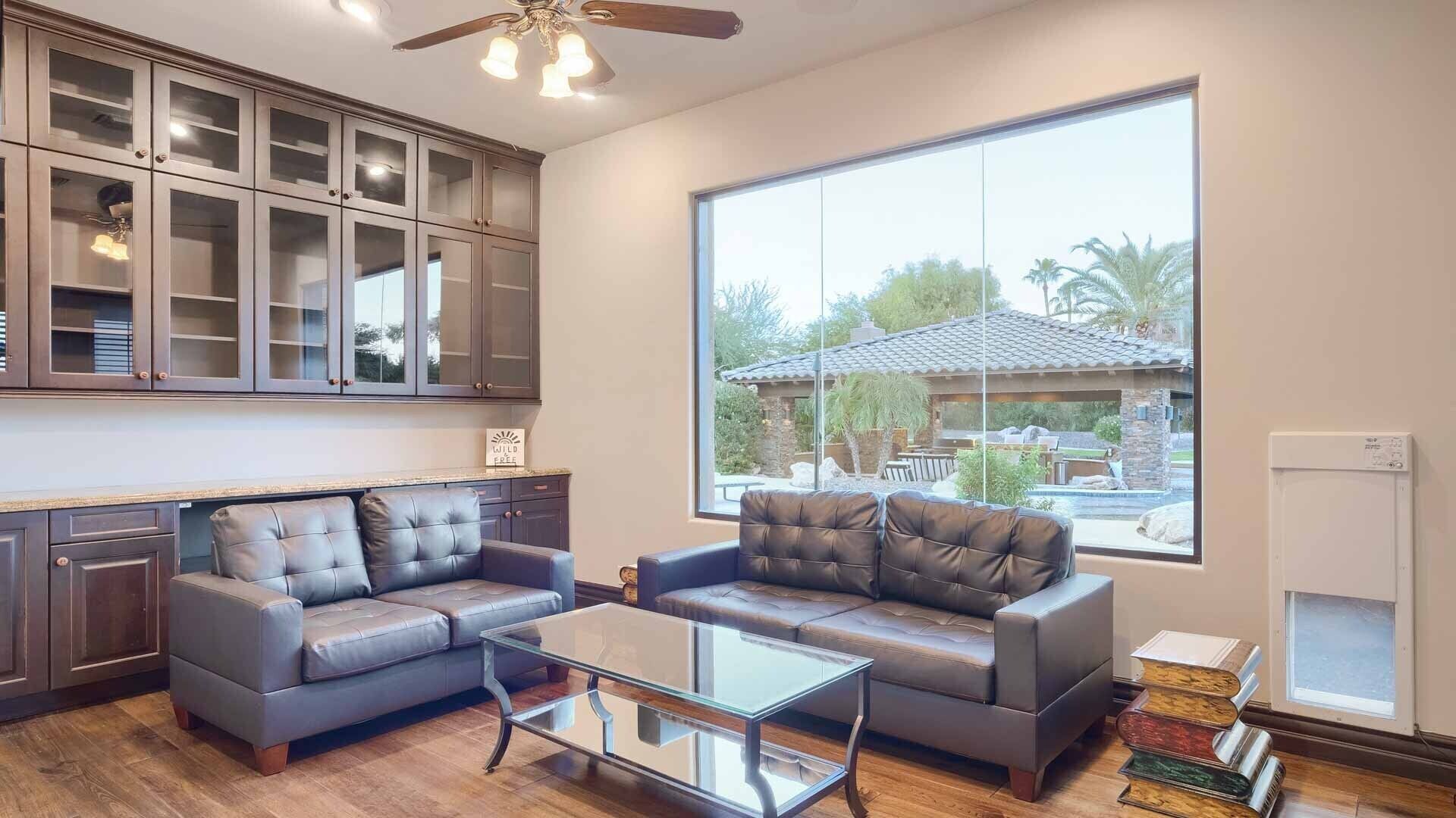 A spacious living room with a large window, leather sofas, a glass coffee table, and a ceiling fan.
