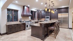 A modern kitchen with dark wood cabinets, a central island with bar stools, stainless steel appliances, and a large window with a view. - 7BR Luxury Oasis w/ Relaxing Lazy River, Basketball, Full Gym & Private Theater (Paradise Valley)