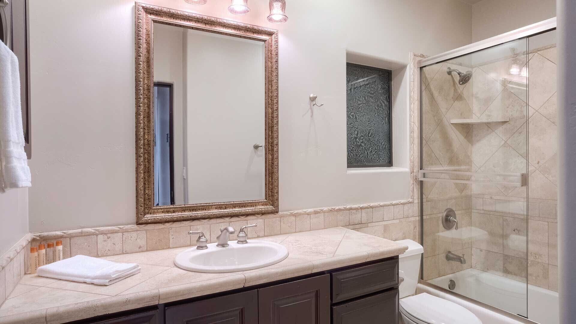 A bathroom with a large mirror, a sink, dark cabinets, a shower, and a small window.