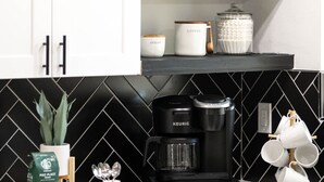 Een moderne keuken met witte kastjes, een zwart betegelde achterwand, een Keurig koffiezetapparaat en een koffiepot op het aanrecht.