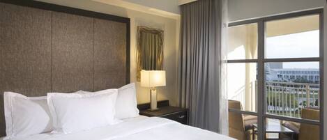 A hotel room with a large bed, a nightstand with a lamp, a view of the city through a balcony with wicker furniture.