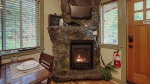 Ein Kamin aus Stein mit einem Fernseher darüber, ein Esstisch mit Tellern und ein Feuer, das brennt.