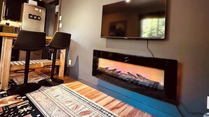 A modern living room with a flat-screen TV mounted on the wall, a built-in fireplace, and two black bar stools.
