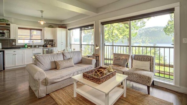 A spacious living room with a sofa, armchair, and coffee table, large windows with a view of greenery, and a kitchen in the background.