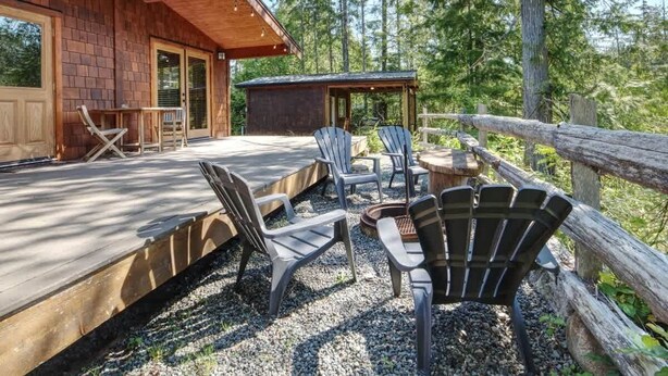 A wooden deck with Adirondack chairs and a fire pit, surrounded by a wooden fence and trees. - Explorers Retreat  Private Sauna - Pet Friendly (Port Renfrew)
