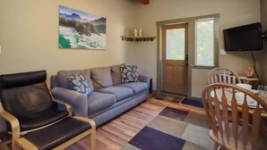 A living room with a sofa, two armchairs, a dining table with chairs, a TV mounted on the wall, and a door leading outside. - Explorers Retreat  Private Sauna - Pet Friendly (Port Renfrew)