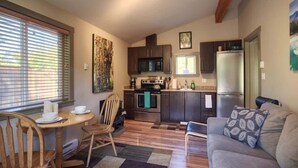 A compact kitchen with dark cabinets, a stainless steel refrigerator, and a microwave. There is a dining area with a wooden table and chairs, and a gray sofa with a patterned pillow. - Explorers Retreat  Private Sauna - Pet Friendly (Port Renfrew)