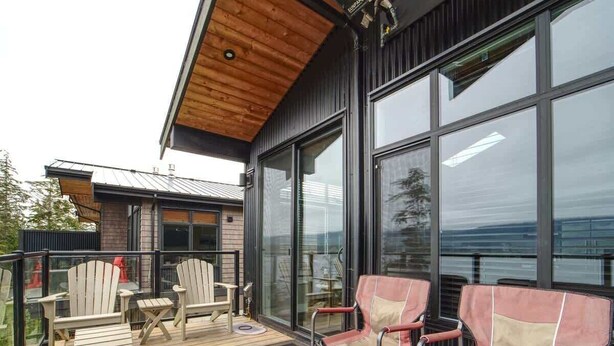 A modern outdoor deck with wooden flooring, two Adirondack chairs, a fire pit, and large windows offering a view of the surrounding landscape. - Amazing Marina & Oceanfront View - Pet Friendly - Sleeps 6 (Port Renfrew)