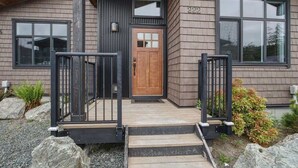 A modern house with a wooden front door, a small porch with a metal railing, and a stone pathway leading to the door. - Amazing Marina & Oceanfront View - Pet Friendly - Sleeps 6 (Port Renfrew)