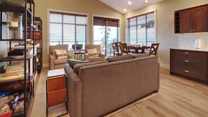 A spacious living room with a sofa, a coffee table, and a dining area with chairs. There is a ceiling fan, large windows with blinds, and a kitchen area with cabinets. - Amazing Marina & Oceanfront View - Pet Friendly - Sleeps 6 (Port Renfrew)