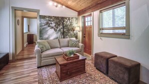 A cozy living room with a wooden ceiling, a beige sofa, a wooden coffee table, and a large window with blinds. - A slice of paradise with Hot Tub - Pet Friendly (Port Renfrew)