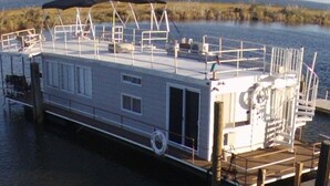 A houseboat docked at a pier with a covered deck area, windows, and a small outdoor seating area. - Awesome retreat on the water with sunrise (Chauvin)
