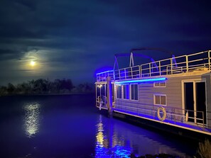 Een woonboot met een overdekt terras en een klein balkon, afgemeerd in de nacht met een weerspiegeling op het water.