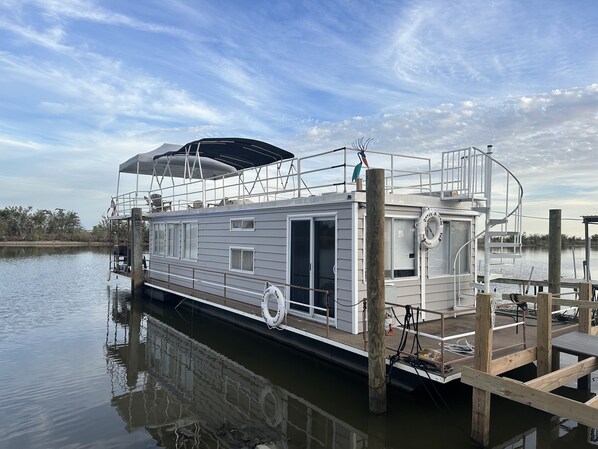 A houseboat docked at a pier with a covered deck area, a lifebuoy, and a small outdoor seating area.