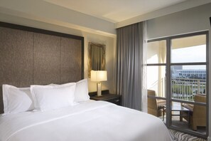 A hotel room with a large bed, a nightstand with a lamp, a view of the city through a balcony with wicker furniture.
