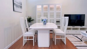 A modern living room with a white dining table, matching chairs, a TV, and a cabinet with glass doors. - Spacious Sunlit 3BR • Center of Downtown Boston • Family Favorite (Boston)