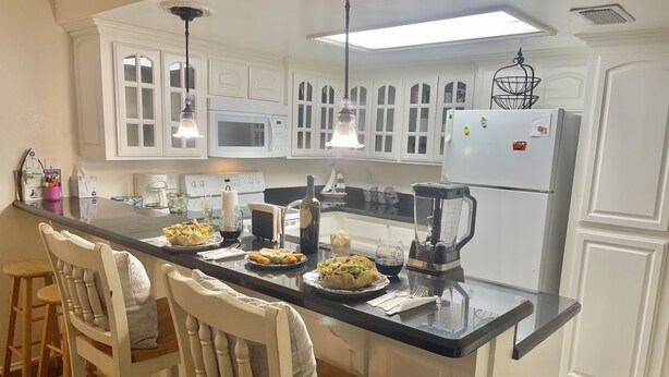 A kitchen with white cabinetry, a black countertop, and a central island with chairs. There are various appliances including a microwave, stove, and refrigerator.