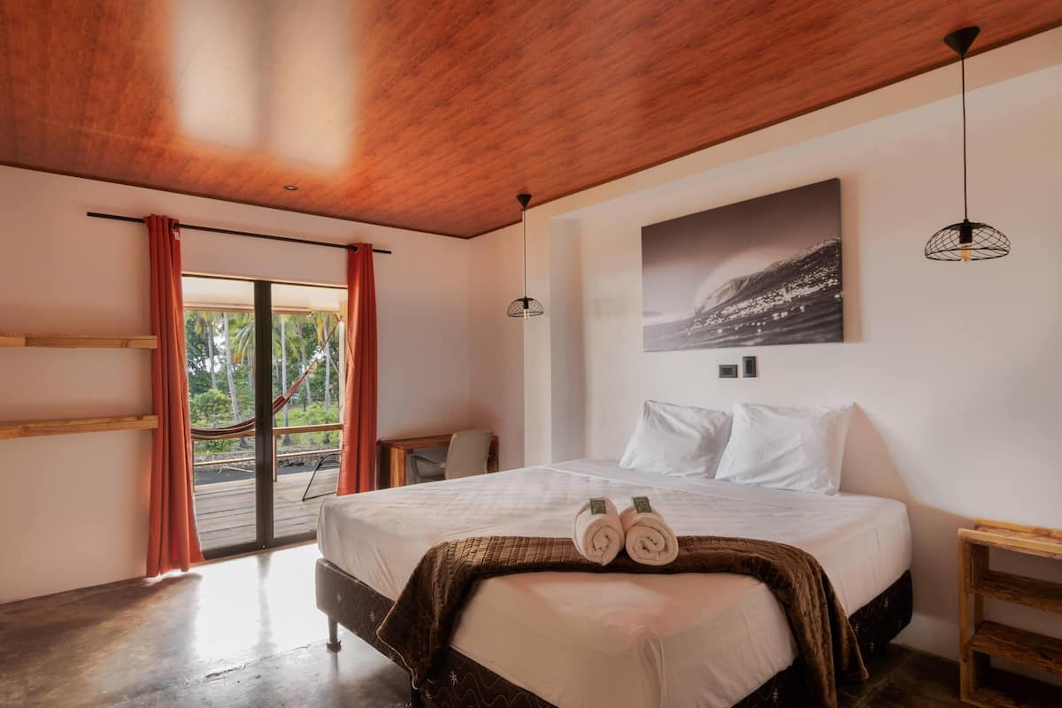 Un dormitorio con cama, techo de madera, ventana con cortinas, un cuadro en la pared y una hamaca afuera.