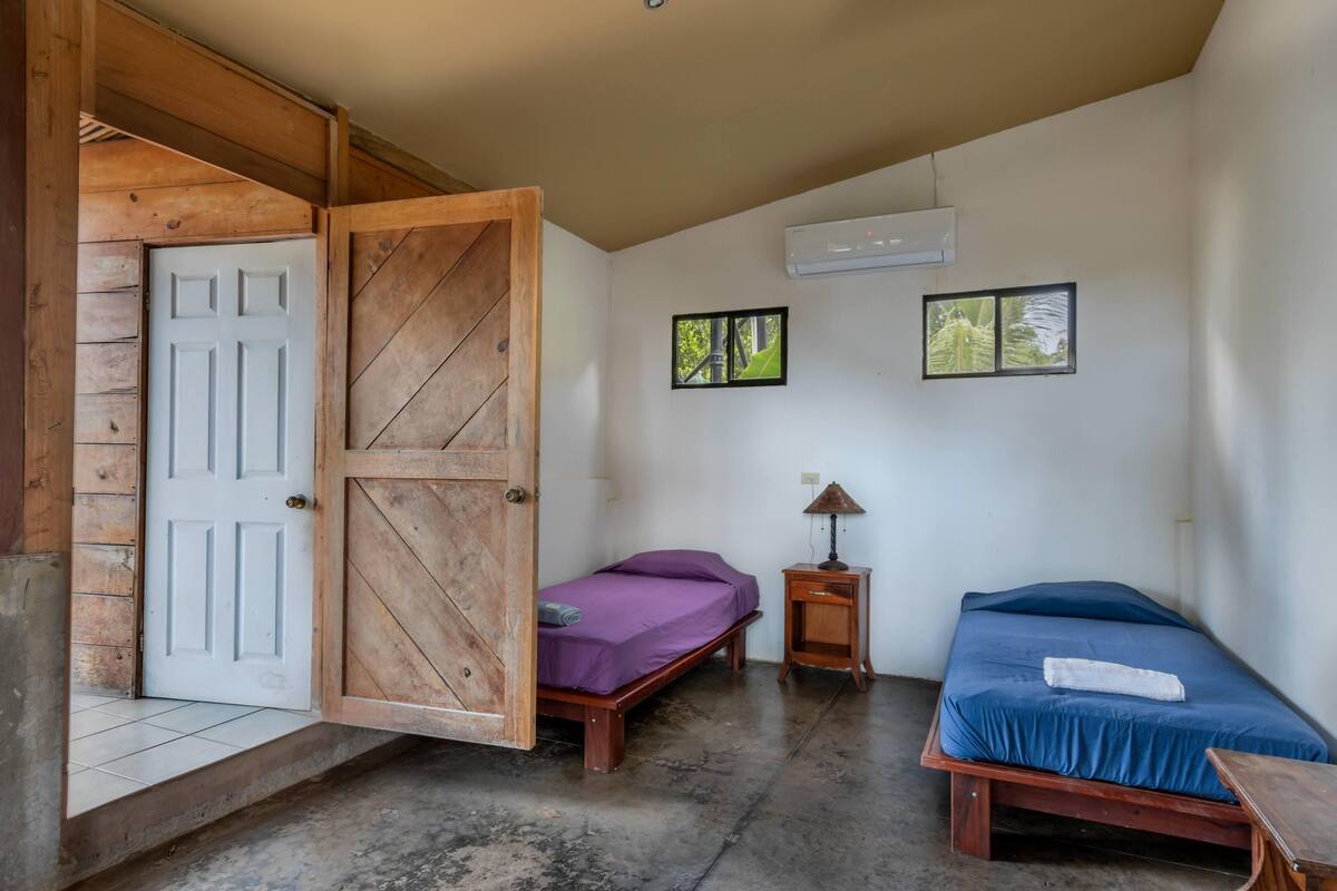 Una habitación con dos camas, una puerta de madera, un aire acondicionado y dos cuadros en la pared.