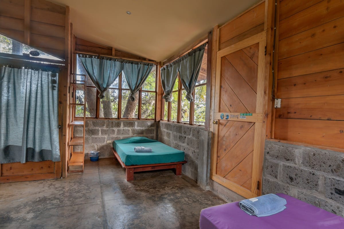 Una habitación pequeña y rústica con una cama individual, una puerta de madera y una ventana con cortinas.
