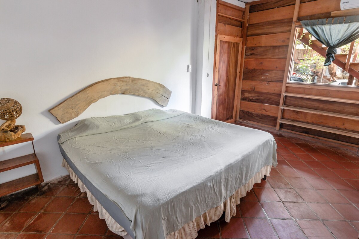 Une chambre avec un lit, une tête de lit en bois, des murs blancs et une fenêtre donnant sur un paysage agréable.
