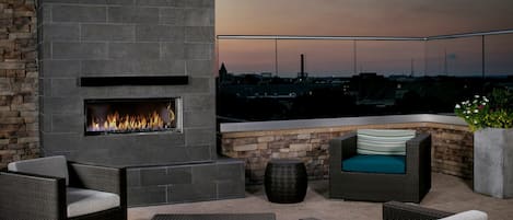 A rooftop patio with a stone fireplace, wicker furniture, and a view of the cityscape at dusk.